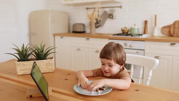 a Little Caucasian Boy Eats Krunchy and Watches Tablet PC at Kitchen Table