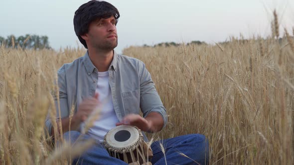 Handsome Man in a Gray Shirt is Sitting in a Wheat Field and Playing a Drum alt