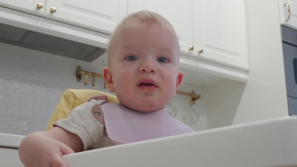 Mother Feeding Oneyearold Baby Boy with a Spoon and a Bowl in the Kitchen at Home alt