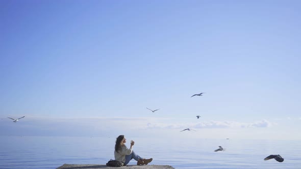 Woman Drinks Coffee Sitting on Sea Pier While Seagulls Fly