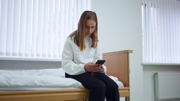 Absorbed Young Woman Messaging Online on Smartphone Sitting in Hospital Ward Indoors alt