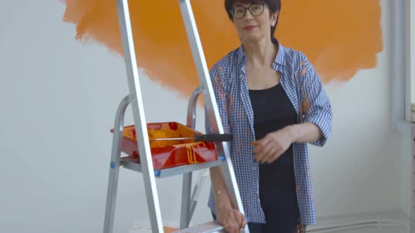 Flat Renovation Concept. Happy Middle-aged Woman Painting White Wall with Paint Roller, Orange Paint alt