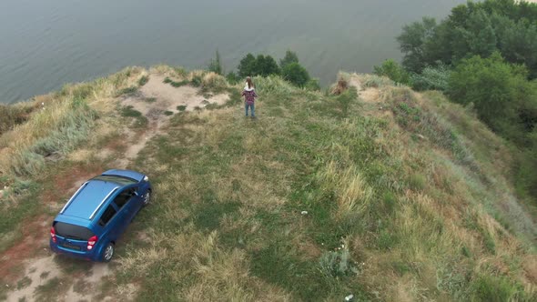 Little Caucasian Girl with Long Hair Sitting on Shoulders of Man Standing on Cliff Over River in alt