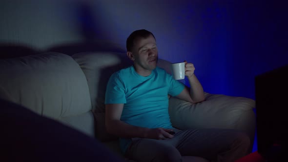 A man with a mug of hot tea sits at the TV late in the evening alt