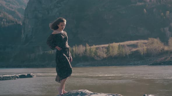 Graceful Woman Stands on Rock at River Against Mountains alt