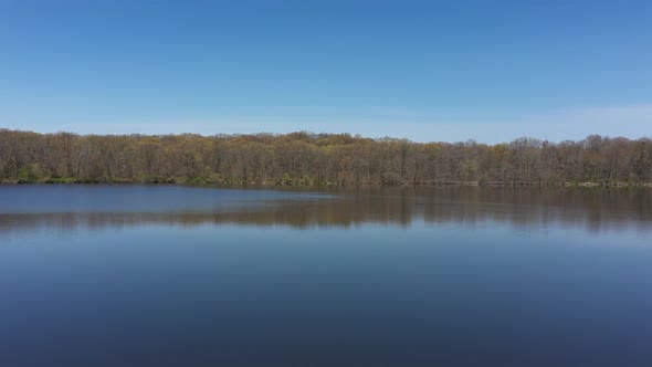 An aerial view of a blue, reflective lake with bare trees lining the lake's edge. The sky is blue & alt