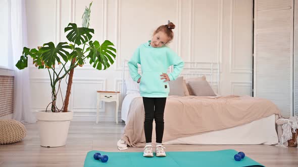 Beautiful Elementary School Girl Performs Sports Exercises at Home on a Mat with Dumbbells in the