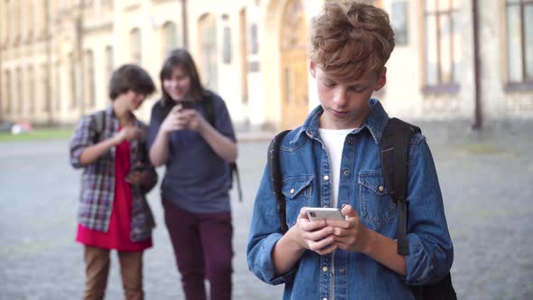 Sad Redhead Caucasian Schoolboy Using Smartphone, Sighing. and Looking Back at Classmates Teasing alt