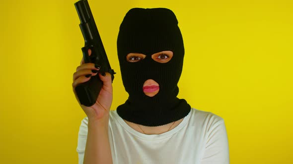 Unrecognizable woman in black balaclava with gun on yellow background. alt