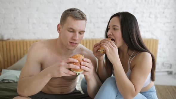 Young Couple Man and Woman Are Eating Hamburgers Together After Workout at Home