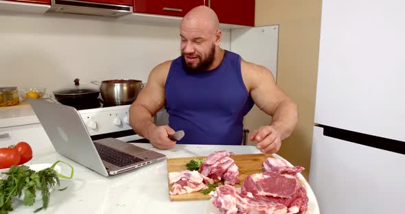 Cheerful Man Is Sitting at Home, Chopping Beef and Viewing Social Nets at Notebook alt