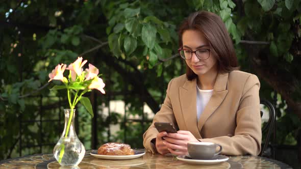 A Young Business Woman Is Sitting in a Cafe and Texting on the Phone. Girl with Coffee and a Bun on alt