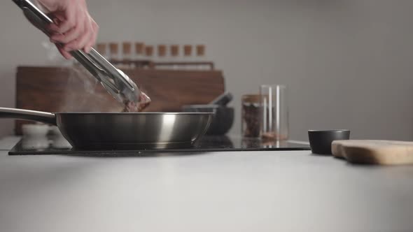 Man Frying Beef Steak Searing Side on Steel Pan on Electric Hob on Kitchen alt