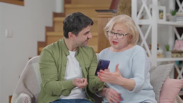 Positive Adult Son Explaining Smartphone App Messaging to Senior Beautiful Mother in Eyeglasses alt