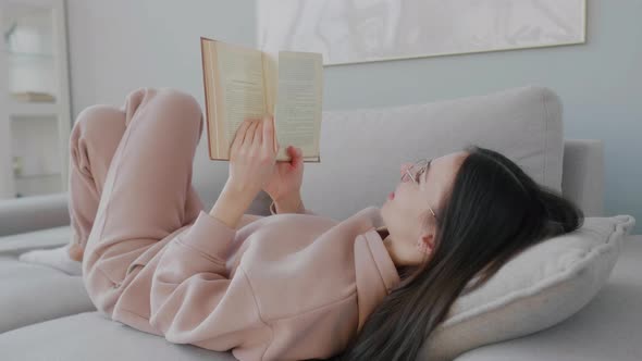 Young Caucasian woman wearing reading glasses lying on sofa and reading book.