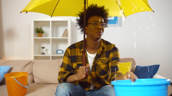 African Guy at Home Under Umbrella Holding Plastic Bucket Collecting Water Falling From Ceiling alt