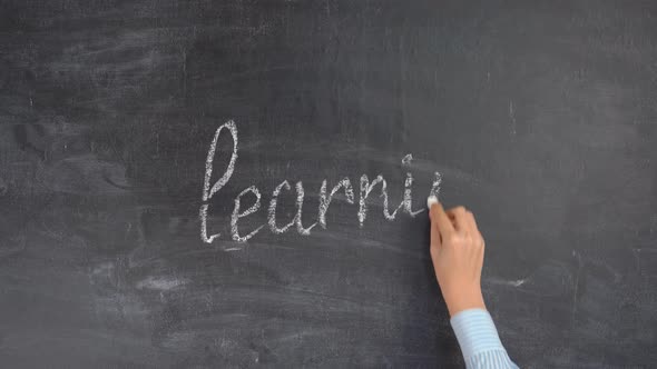 The hand of a female teacher writes the word LEARNING on a black chalkboard with white chalk alt