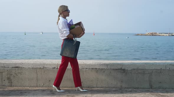 Sad Elegant Businesswoman Walking in Slow Motion with Box Looking at Mediterranean Sea at Background alt