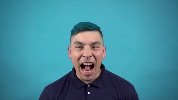 A Young Man in Glasses with Blue Hair Screams in Anger. Emotional Man Screaming on a Blue Background alt