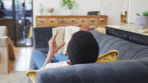 Happy african american woman lying on sofa in living room, reading book alt