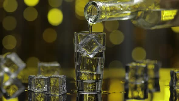 Barman Pour Frozen Vodka From Bottle Into Shot Glass. Ice Cubes Against Shiny Gold Party Background alt