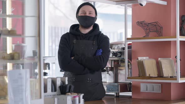 Positive Male Barista in Coronavirus Face Mask Crossing Hands Looking at Camera Standing at Cafe alt