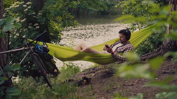 Man Travels on Bicycle Relaxing in Green Hammock Surfing Internet on Smartphone Listening Music on alt