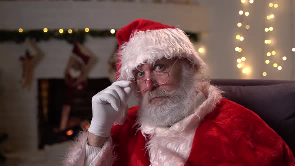Portrait of Funny Santa Claus in Glasses, Sitting in His Rocking Chair and Puts His Finger To His