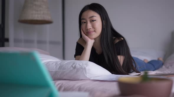 Happy Smiling Asian Young Woman Looking at Camera Lying on Comfortable Bed in Home Office alt