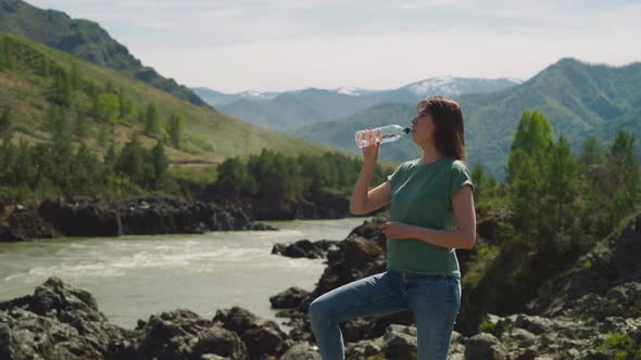Tired Woman Drinks Water and Looks Around on River Bank alt