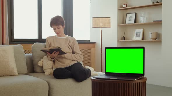 A Laptop with a Green Screen Stands on a Table Against of a Woman Reading a Book alt