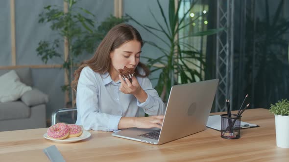 Young Businesswoman Eating a Glazed Donut While Working on Laptop in Office alt