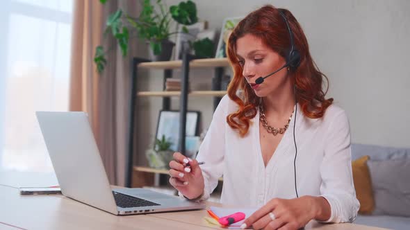 Caucasian Female Distant Worker Wears Headphones Video Calling Makes Notes alt