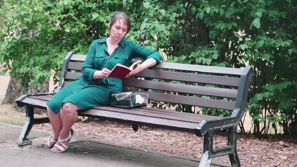 An adult woman is reading an interesting book while sitting on a park bench