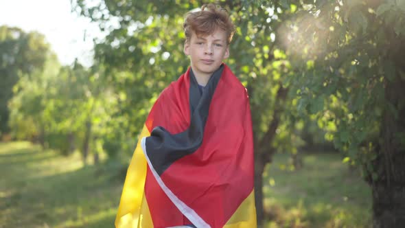 Portrait of Teenage Caucasian Redhead Boy with Curly Hair Wrapped in German Flag Looking at Camera alt