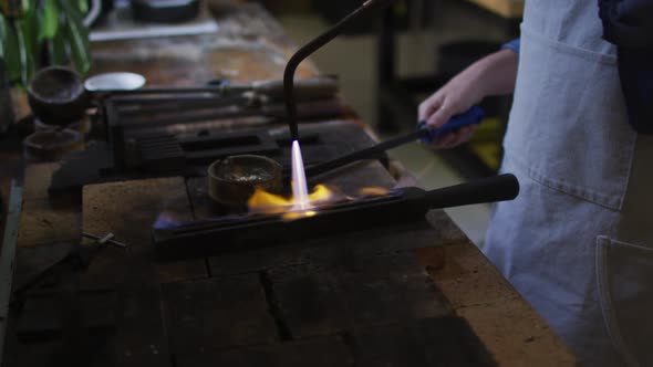 Midsection of caucasian female jeweller in workshop wearing apron, using gas burner alt