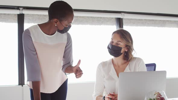 Two diverse female colleagues wearing face masks looking at laptop and discussing in office alt