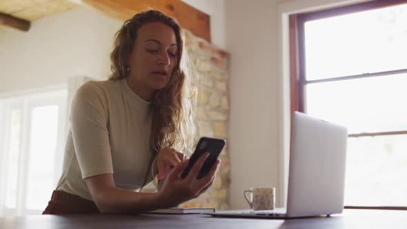 Caucasian woman working from home, sitting at desk using smartphone and laptop alt