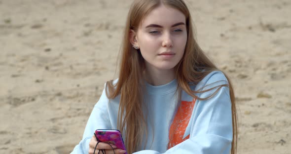 Young Beautiful Girl Sitting on the Beach and Holding a Telephone in her Hands