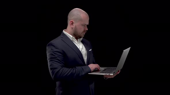 Bald Bearded Handsome Man Is Standing in White Shirt and Dark Blue Suit Typing on Laptop. Black alt