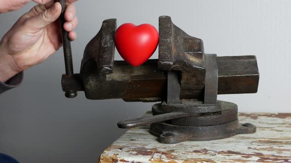 Heart in a vice. Man's hand rotates handle of rusty grip vice and red heart is squeezed