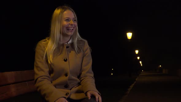 A Young Beautiful Woman Sits on a Bench and Celebrates in an Urban Area at Night alt