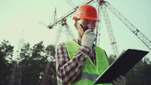 Electrical Equipment Worker Near High Voltage Tower Using Walkie Talkie and Clipboard alt