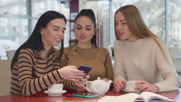 Close-up of Three Caucasian Female Friends Looking at Smartphone Screen in Restaurant and Talking alt