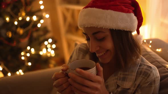Woman in Pajama and Red Santa Hat Sitting on Couch Watching TV and Drinking Hot Chocolate From Mug alt