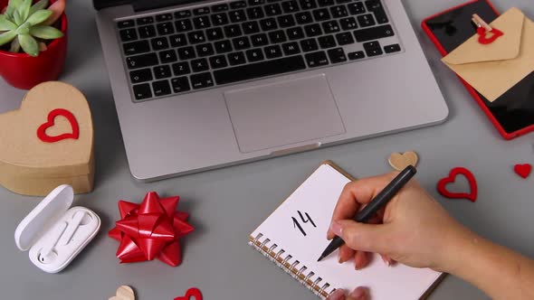 Somebody writing 14 FEBRUARY in a notebook near laptop alt