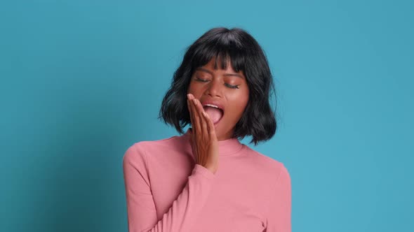 Dark Skinned Woman Yawns with Her Mouth Wide Open on a Background of Blue Wall alt