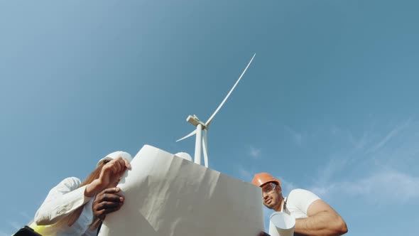 Four Multi Ethnic Partners in Safety Helmets Studying Wind Turbine Blueprints alt
