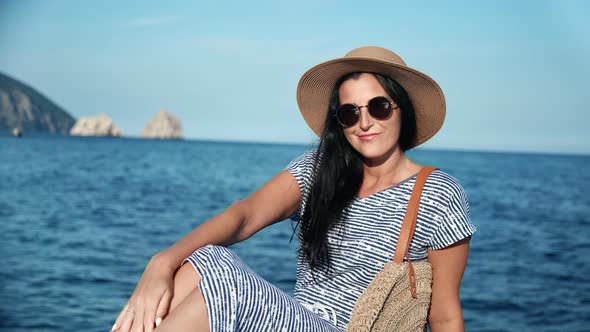 Sexy Female in Sunglasses and Straw Hat Sitting at Seashore alt