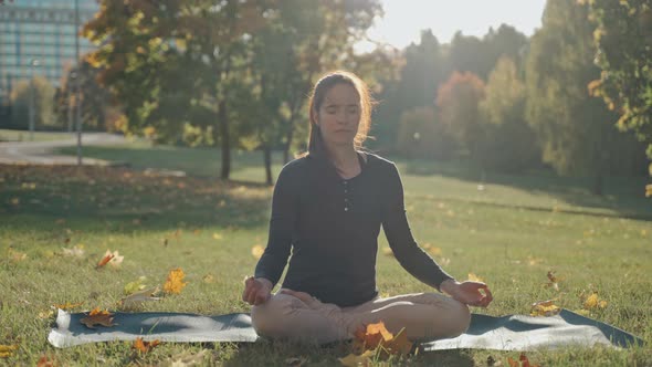 Young Woman Sitting on Mat in Lotus Position in City Park on Sunny Autumn Day alt